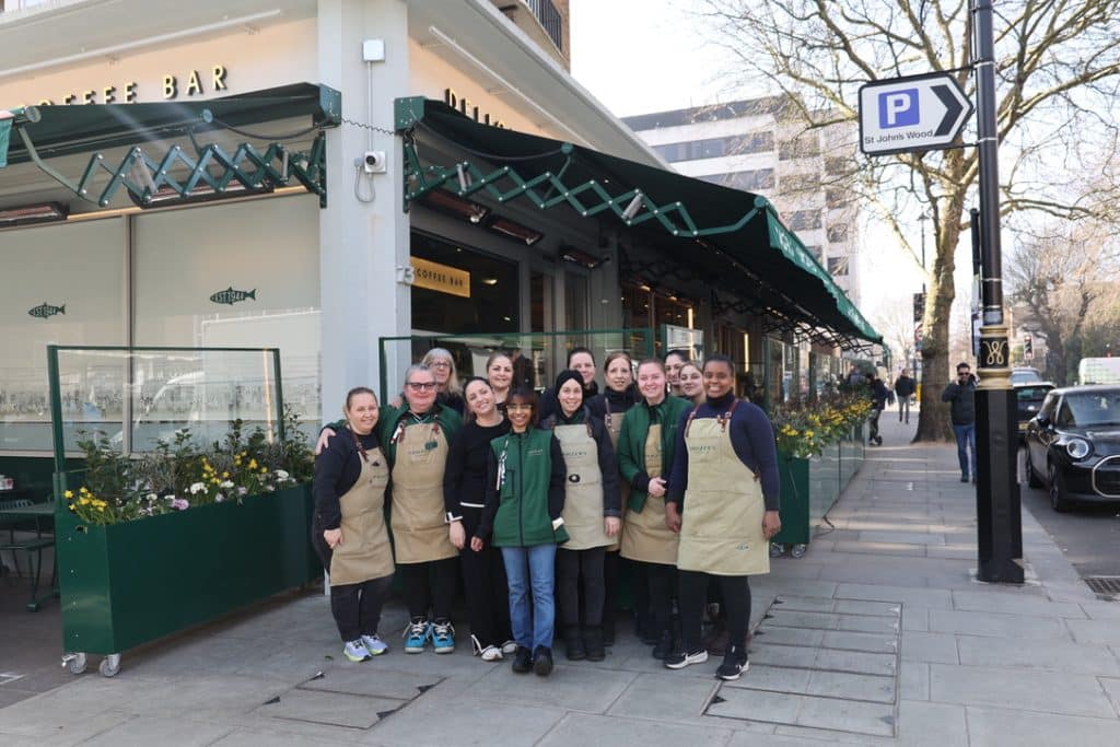 A group of women from the Panzer’s team stand together outside the shop, smiling at the camera. They are dressed in Panzer’s uniforms, including beige aprons and green jackets, with the store’s signage and flower arrangements visible in the background.