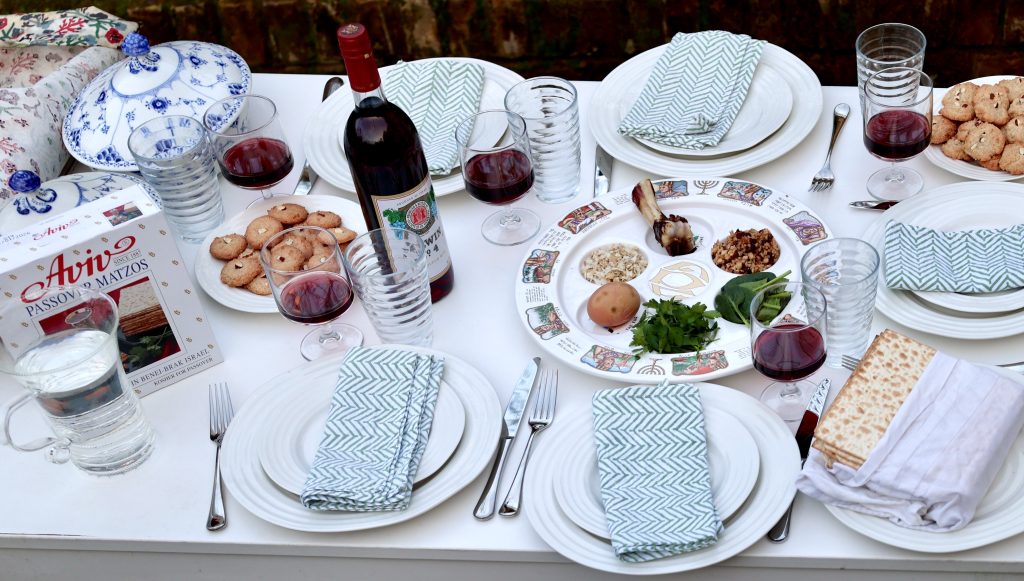 This image shows a beautifully set Passover Seder table. A Seder plate is at the center, featuring traditional symbolic foods such as a shank bone, an egg, bitter herbs, charoset, and parsley. Surrounding the Seder plate are plates with folded napkins, glasses filled with red wine, and a bottle of kosher wine. A box of Aviv Passover matzos is present, along with a covered stack of matzo on one of the plates. The table also includes plates of cookies, elegant blue-and-white covered serving dishes, and glassware. The setting is outdoors, with a white tablecloth creating a clean and festive presentation.